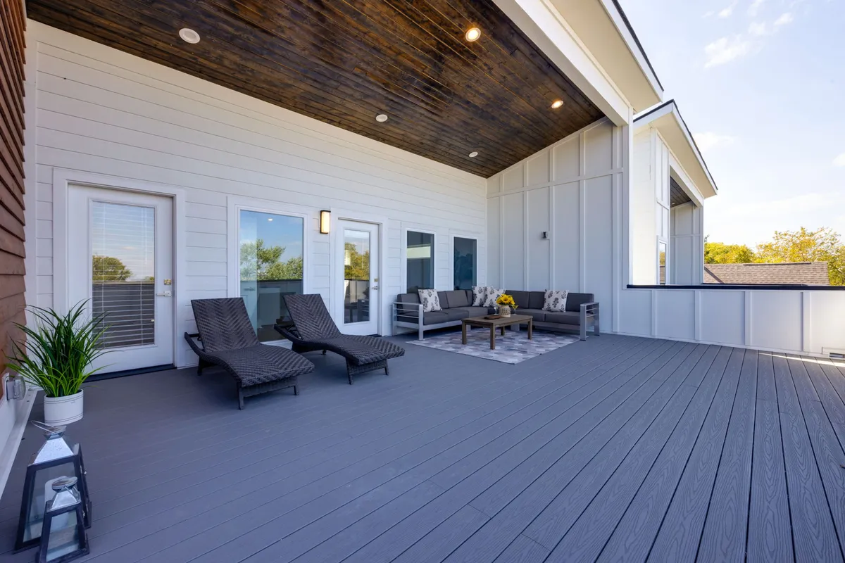 A tranquil rooftop deck featuring comfortable outdoor seating with decorative pillows, a sunflower centerpiece on the coffee table, lantern, and deck flooring. The setup conveys a cozy escape with open sky views, ideal for relaxing or entertaining.