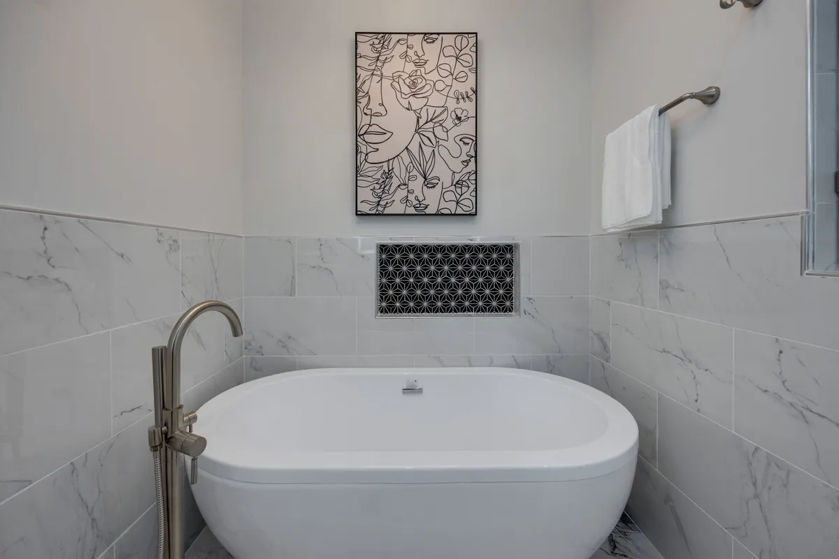 A spa-inspired bathroom featuring a freestanding bathtub, marble tile, and artwork on the wall. Soft, ambient lighting contributes to a tranquil, luxurious bathing experience.