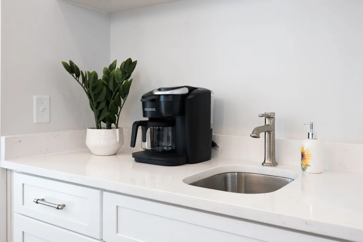 A close-up of the coffee maker, plant, and kitchen sink highlights the thoughtful amenities provided. The scene feels cozy and homey, suggesting the ease of starting the day with a cup of coffee, in line with the mention of the Keurig Duo and kitchen starter kit.