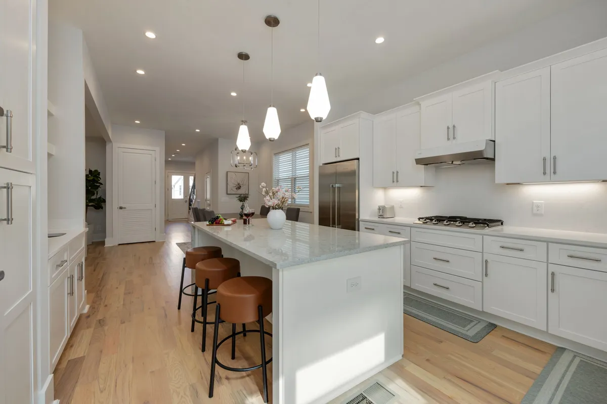 The image showcases a modern, sunlit kitchen with a spacious island, elegant bar stools, high-end appliances, sleek cabinetry, and a decorative vase. The layout invites entertaining, with plenty of space for cooking, gathering, and socializing.