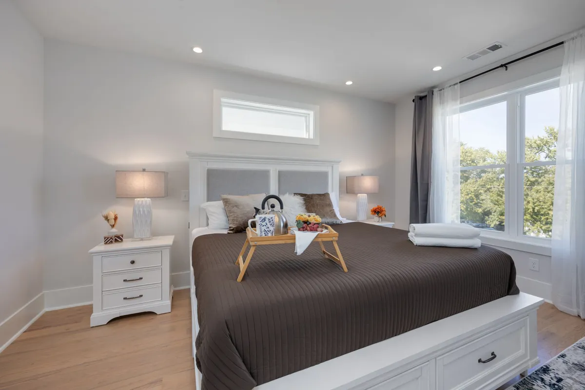 A cozy bedroom with a bed layered in a brown quilted cover, a decorative tray with fruit and a drink, lamps on nightstands, and large windows with curtains providing soft natural light.