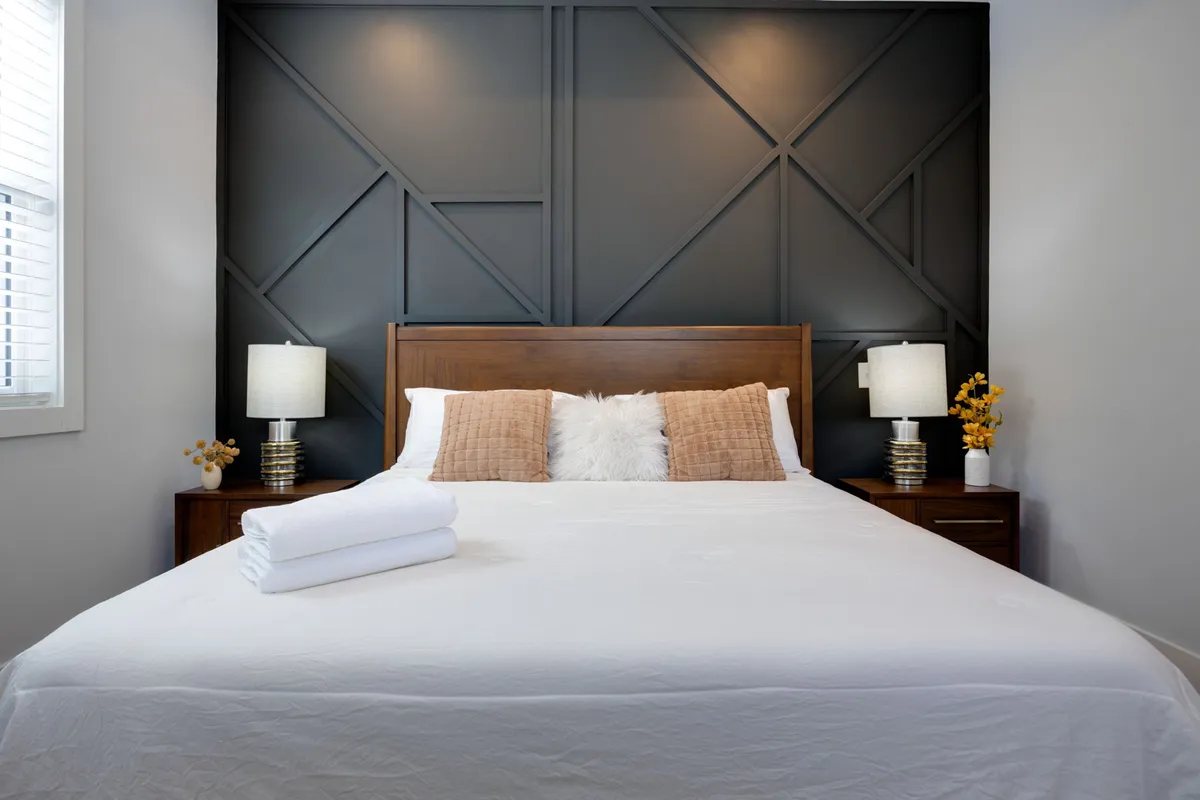 A serene bedroom with a bed dressed in crisp white linens, decorative pillows, geometric accent wall, and table lamps on nightstands. Natural light filters softly through a window, creating a calming and restful atmosphere.