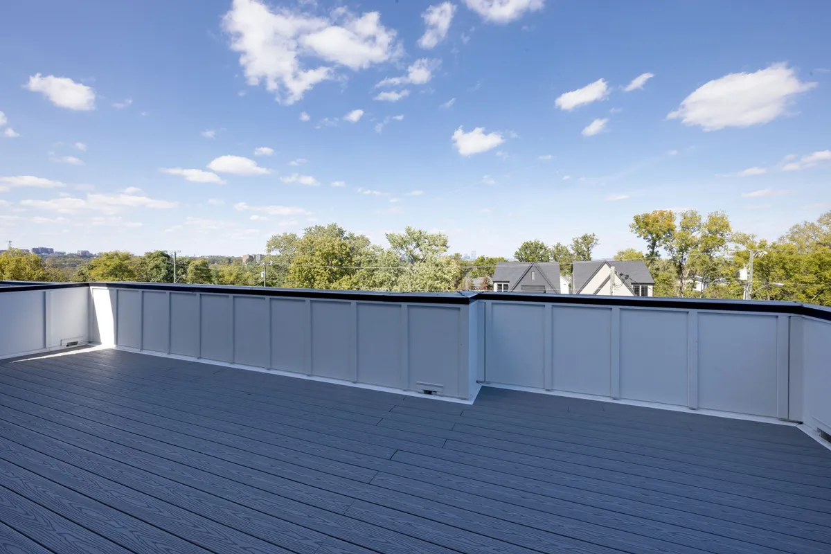 A spacious rooftop deck with comfortable seating, surrounded by greenery and treetops under a vivid blue sky. The scene is tranquil, inviting relaxation or social gatherings, and the height suggests elevated views of the surrounding area.